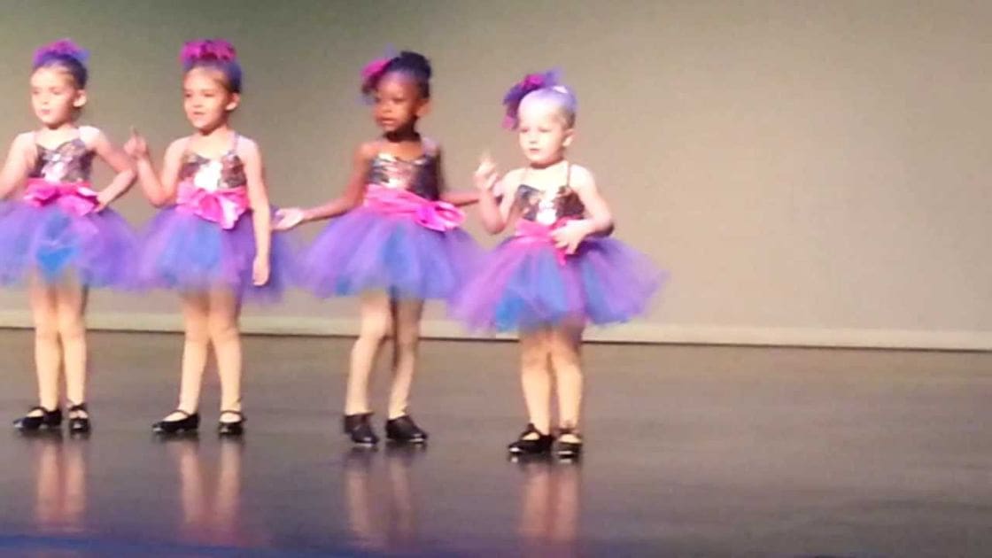 A group of young female children performing on an indoor stage, likely at a dance recital; they wear tutu dresses with various colors and patterns.