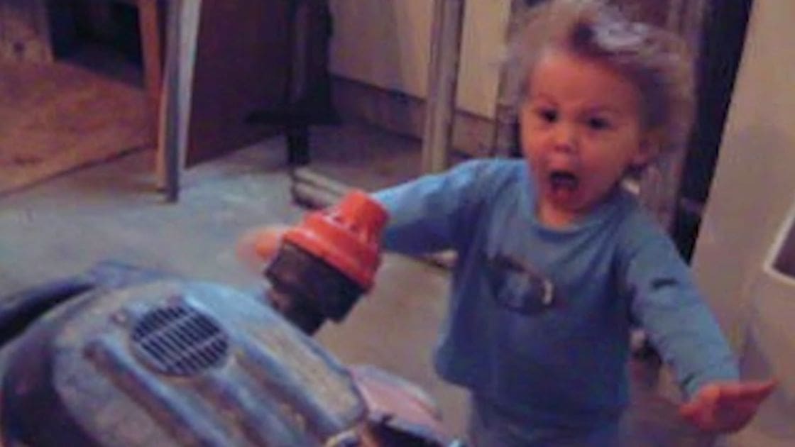 A toddler wearing a light blue long-sleeved shirt with a cartoon graphic, standing indoors near a metallic ride-on toy vehicle.