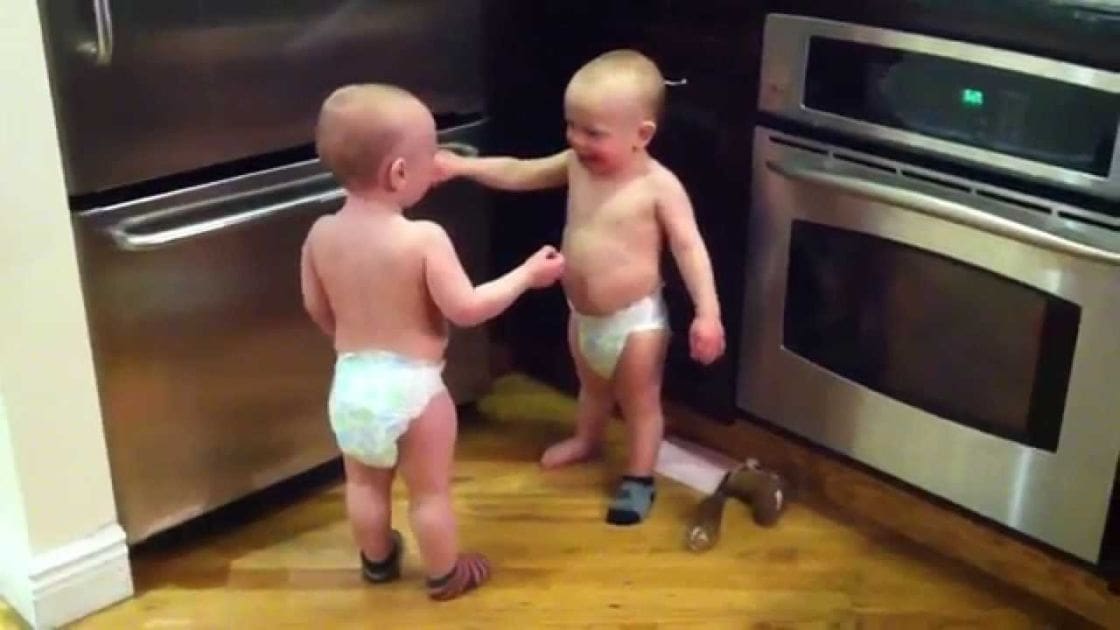 Two toddlers in a kitchen setting with stainless steel appliances, wooden flooring, and a small toy or sandal on the floor.