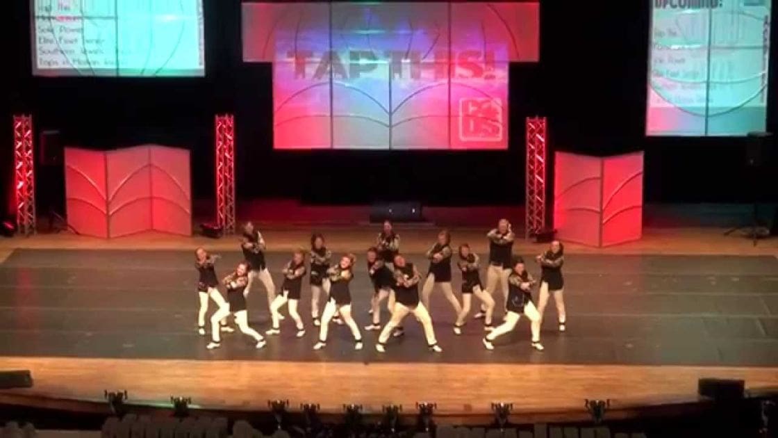 A dance group performing at an indoor event called 'TAP 1153' in a large auditorium, wearing black outfits and white tights on stage under bright indoor lighting.
