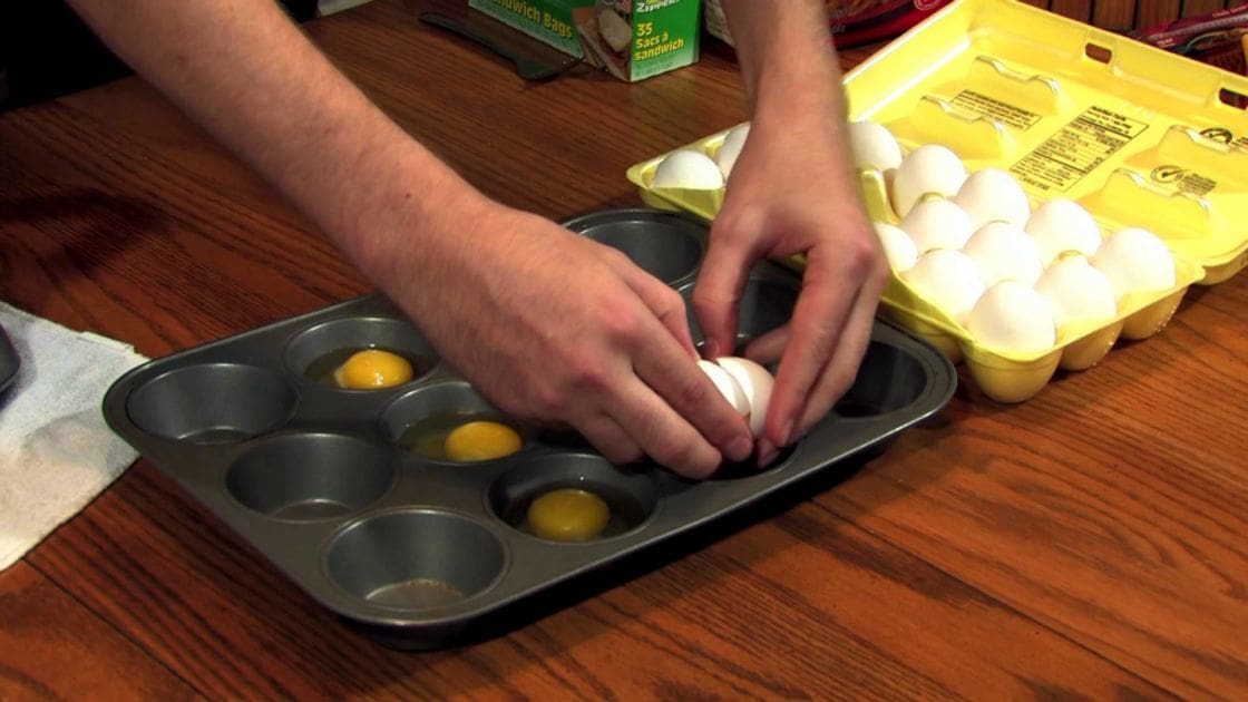 Adult male wearing a dark shirt, preparing eggs in a kitchen with visible cracked eggs and 'Sandwich Bags' box.