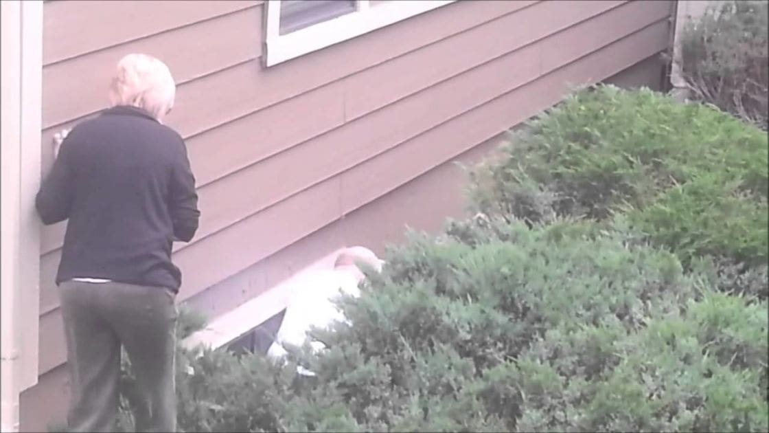 An older adult male stands outside a residential building with beige siding, surrounded by greenery under soft overcast light.