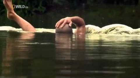 A young adult male in dark-colored swim trunks near a body of water with vegetation and rocks, possibly late afternoon lighting.