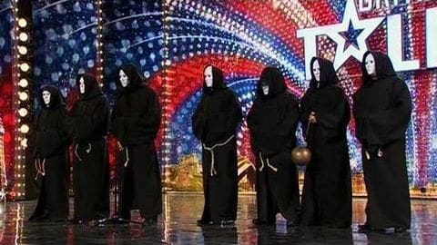 Young adults in black robes with white masks stand on a brightly lit stage resembling 'American Idol' with a backdrop with red, white, and blue colors.