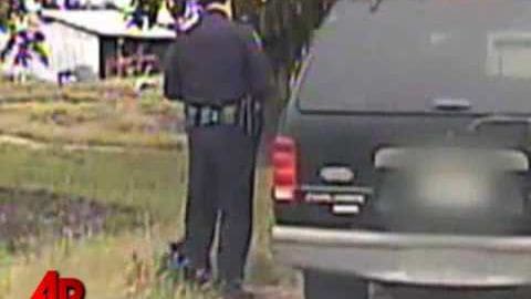 An adult male police officer in a dark blue uniform standing near a gray pickup truck outdoors, with grass and trees visible in the background.