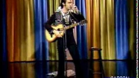 An adult male performer wearing a black and white patterned shirt, dark pants, sneakers, holding an acoustic guitar on stage at a comedy club with blue, orange, yellow, and red vertical striped backdrop.