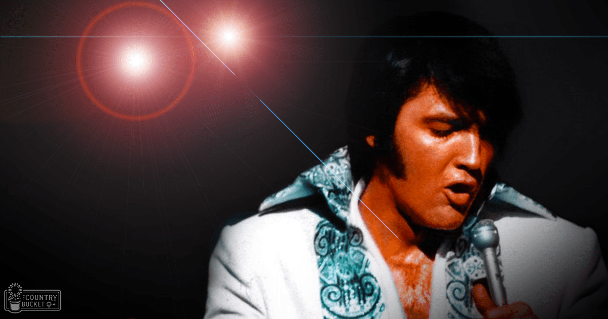 An adult male singer in a white suit with blue embroidery performs on stage under red lights, suggesting a concert setting.