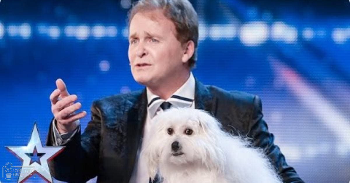 A middle-aged male judge in a dark blazer over a white shirt and black tie on stage with a Maltese dog in the background; indoor venue lighting suggests a talent show setting.