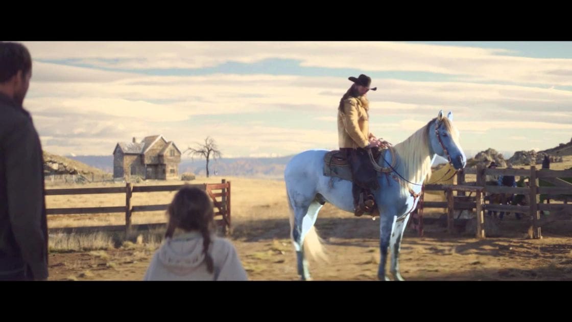 A cowboy rides a white horse with gray markings through an open rural landscape, partly cloudy sky and distant mountains visible.