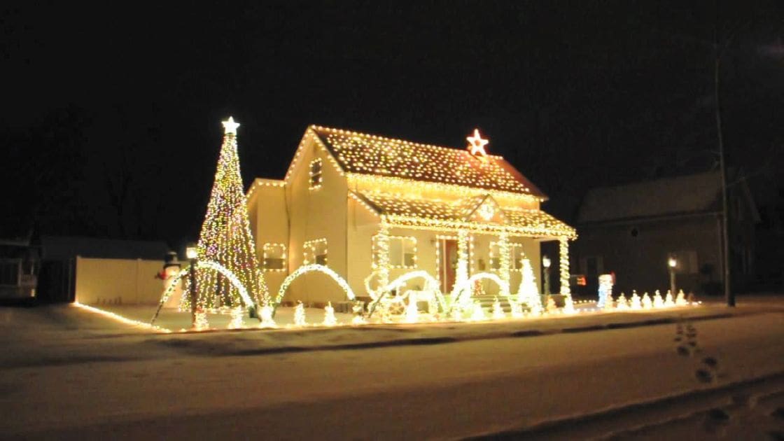 A snowy winter scene with a house decorated for Christmas, with numerous white and yellow lights strung across its roofline and walls. A large illuminated Christmas tree stands to the left of the house, adorned with smaller lights and topped with a star.