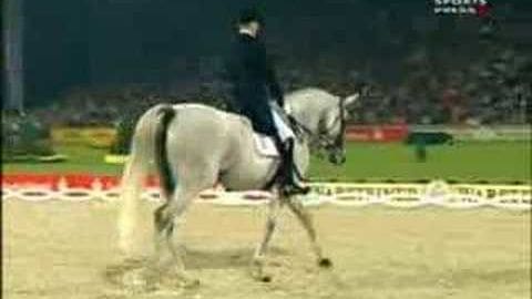 An adult male rider in a dark patterned riding jacket and black boots, on an Arabian horse with white coat and dark mane and tail, inside an indoor equestrian arena with spectators.