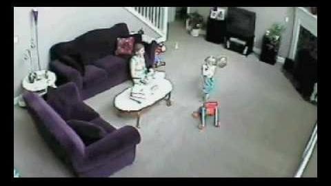 Two individuals in an indoor living room scene with a dark purple sofa, red patterned pillows, white coffee tables, and a child's bicycle near them. The setting appears to be during daytime.