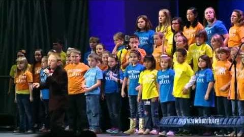 Children in an indoor performance setting, wearing 'Worship Team' t-shirts, standing on a stage with curtains and lighting.