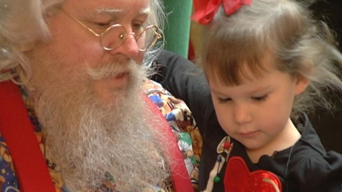 An older adult male singer wearing a star and moon patterned shirt with a red sash, accompanied by a toddler girl in a black heart top, both indoors during what appears to be a festive holiday event.