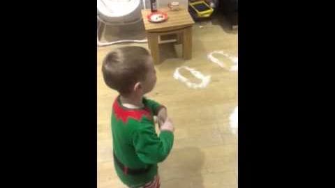 A toddler wearing a festive green sweater with red accents in an indoor residential space with tiled flooring.