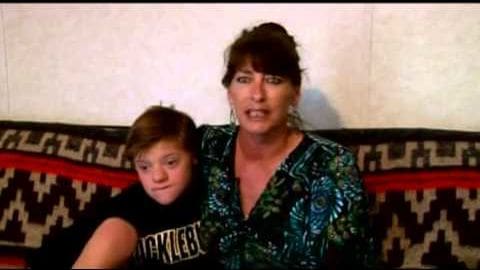 A middle-aged female and a young boy seated indoors, wearing patterned clothing; red and black sofa in background with even natural lighting.