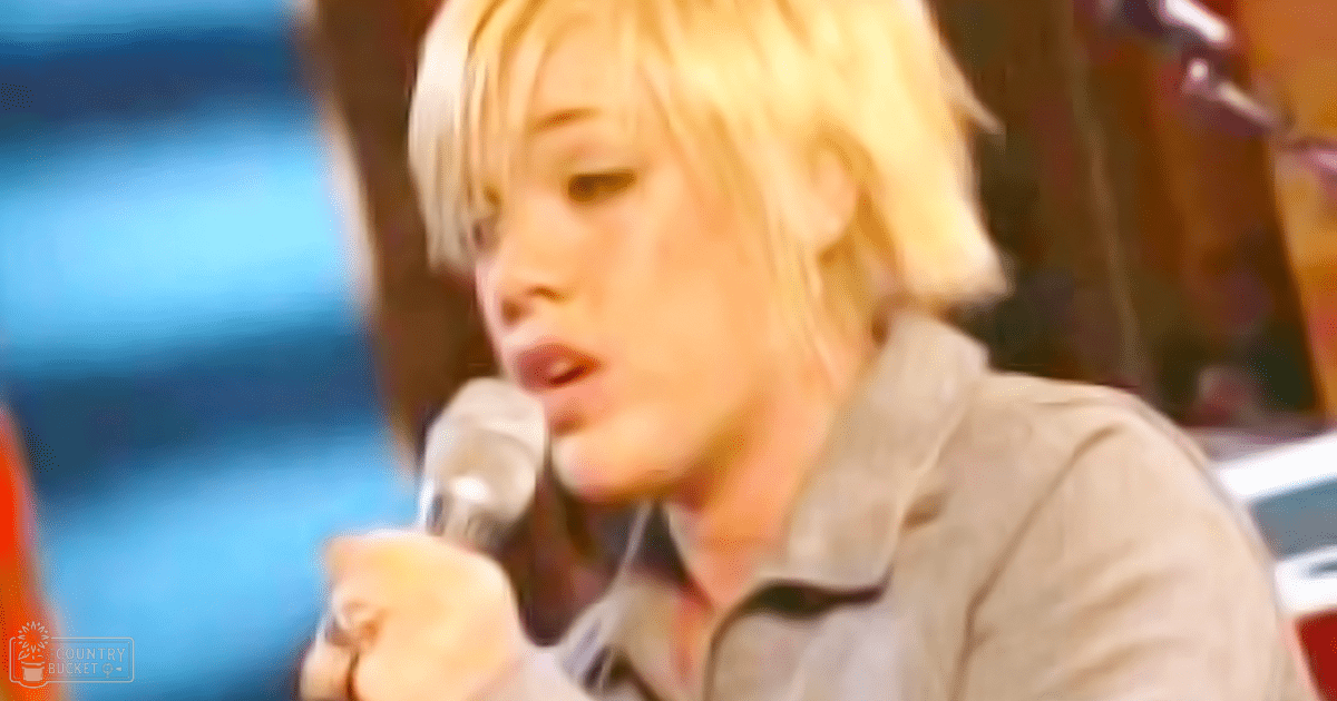 A young adult male singer in a modern casual light gray button-down shirt, holding a microphone on stage with blurred background elements suggesting an indoor venue.