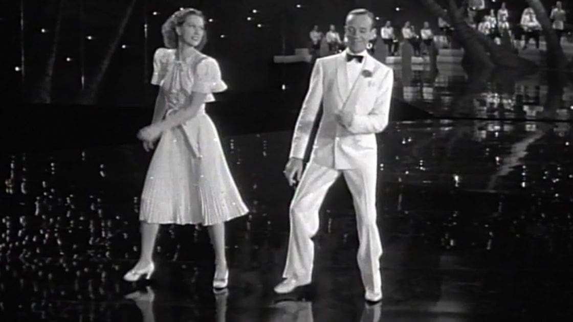 A classic dance scene from a film set on a stage or ballroom floor with dancers in formal attire under artificial evening lighting.