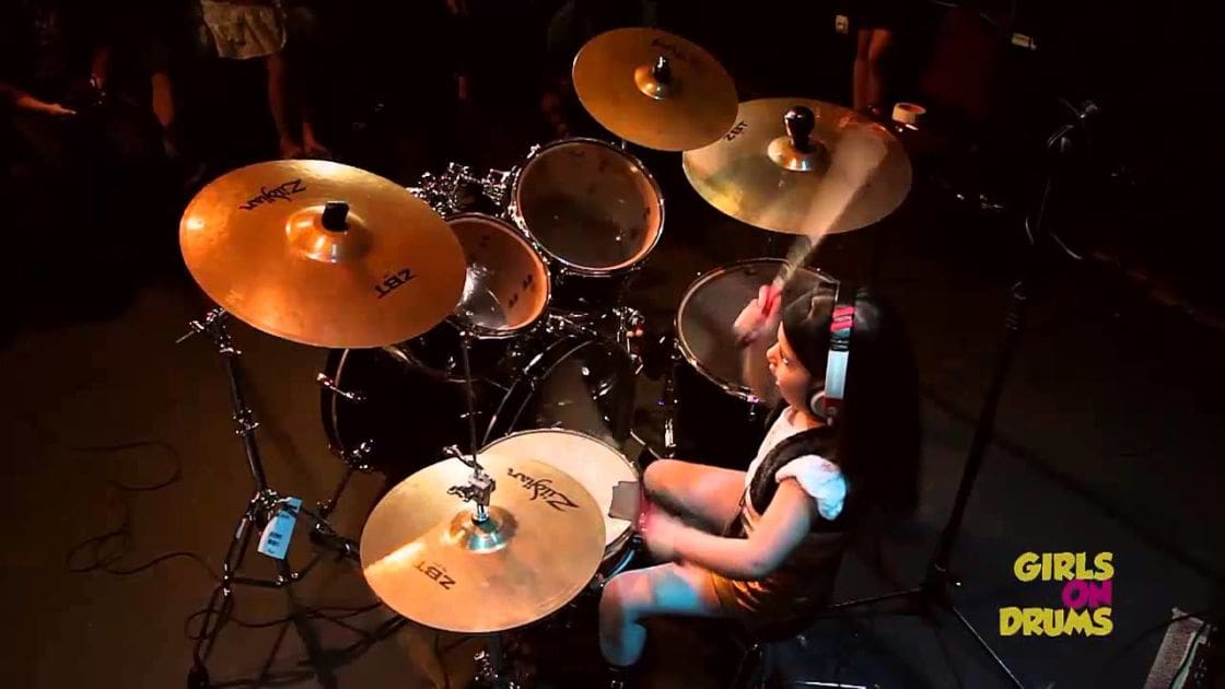 A young adult female drummer in a form-fitting black and white outfit plays drums with a cymbal setup, microphones, and wires visible around her.