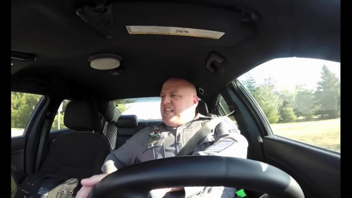 A middle-aged male wearing a dark-colored law enforcement or security uniform with patches and a tactical vest, driving a car in an outdoor setting with bright daylight and greenery visible through the windows.