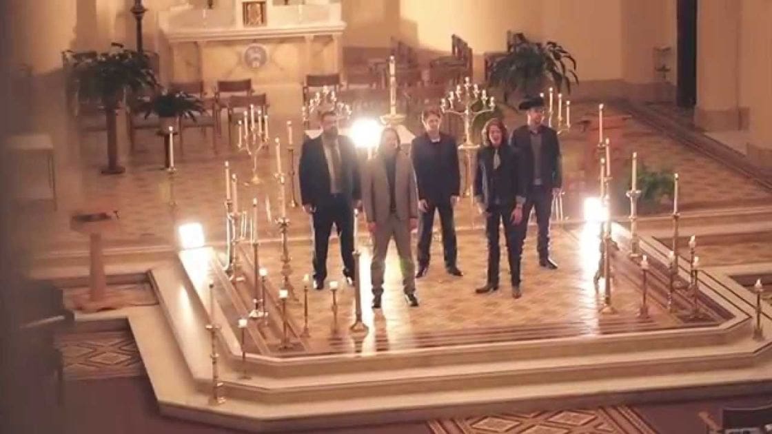 A group of young adults in a church-like indoor setting with polished wood floors and decorative elements during an evening event.