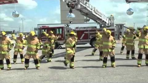 Young adult male firefighters in modern yellow and black reflective uniforms dancing in front of a fire station with red and white fire trucks parked behind them under clear skies.