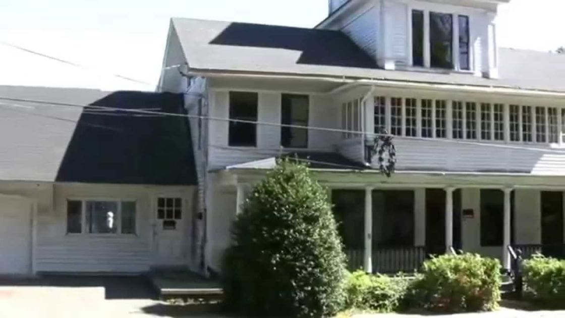 A white two-story house with a porch in an urban residential area under clear daytime skies.