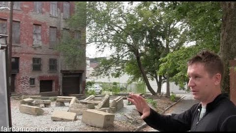 Young adult male wearing a black, relaxed-fit jacket and collared shirt under a historical urban setting with old brick buildings, stone remnants, and trees suggesting spring or summer.