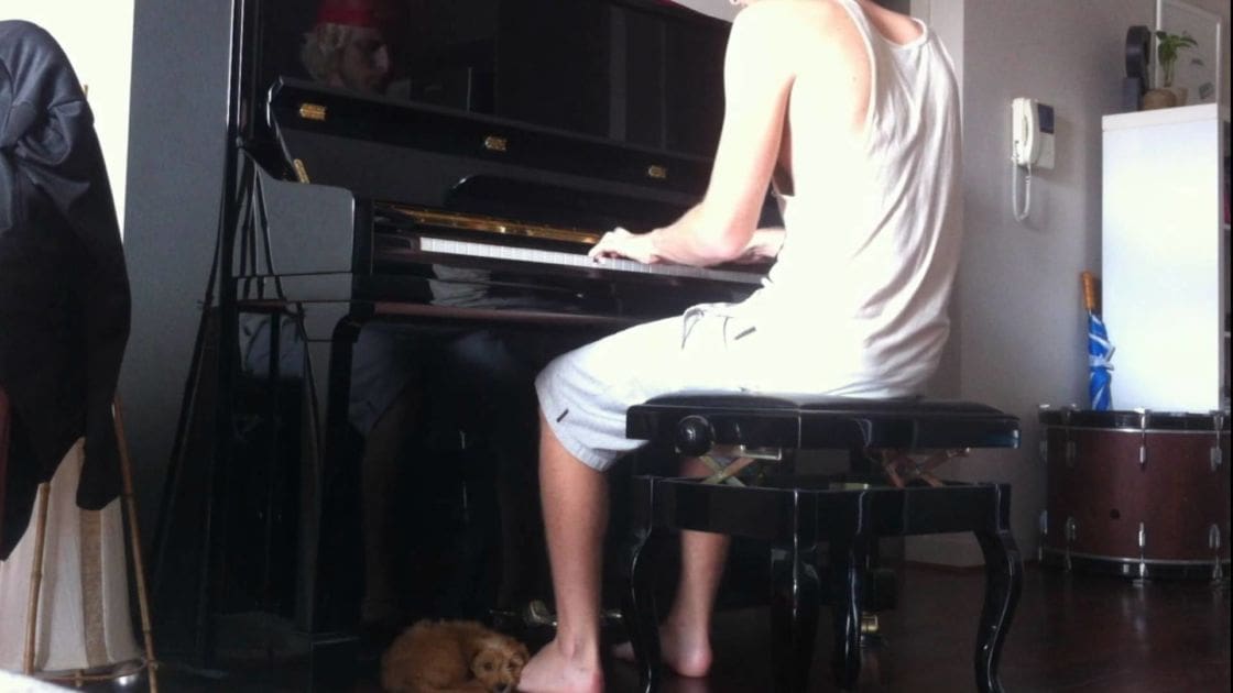 A young adult male plays piano in a home setting with a small dog nearby.