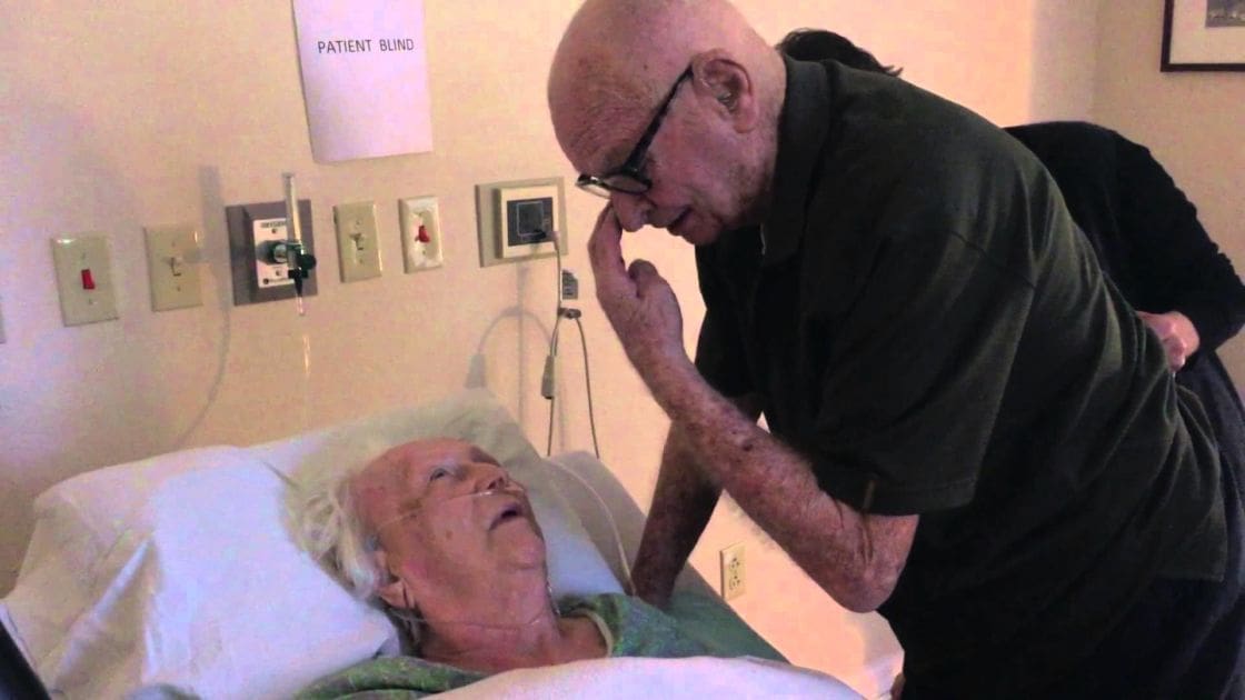 An older adult female in a hospital gown lies in bed, an older male stands beside her with glasses on his nose; the setting is indoors with artificial lighting and white walls.