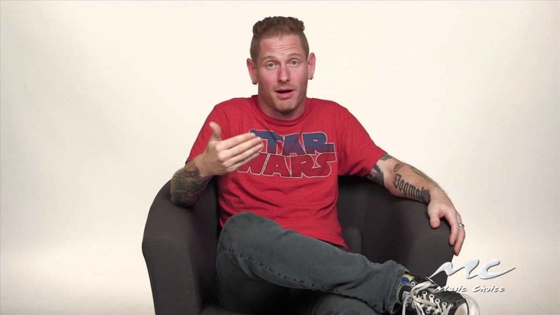 A young adult male wearing a 'Star Wars' logo red T-shirt, dark gray jeans, and black-and-white Converse-style sneakers is seated in an indoor interview setting with plain light-colored background.