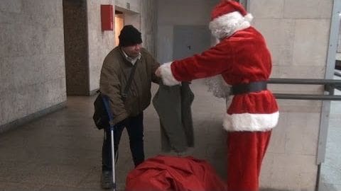 A middle-aged man dressed as Santa Claus stands next to an older adult male, both indoors with concrete walls and a railing; season appears cold.