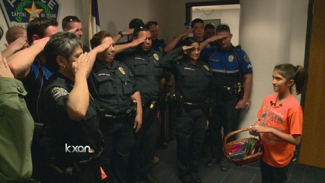 A young girl in an orange t-shirt salutes alongside police officers indoors at the Capital of Texas building.