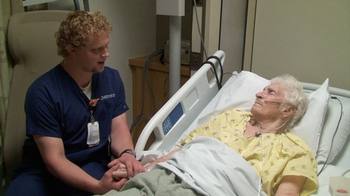 Healthcare professional in blue scrubs interacting with older adult patient in a hospital room setting.