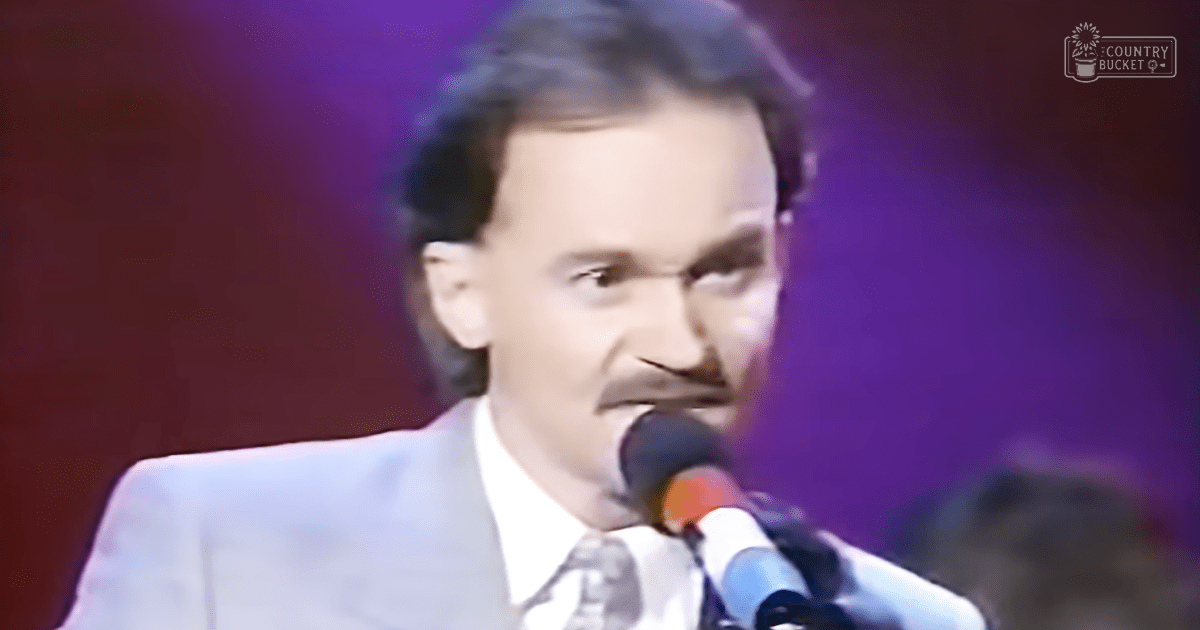 An adult male singer in a light-colored suit jacket with a patterned tie, performing on an indoor stage illuminated by purple lights.