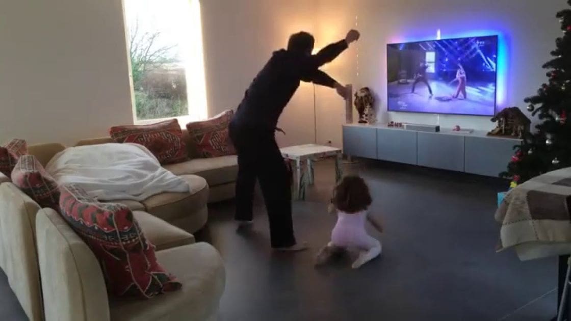 A living room decorated for Christmas with a man dancing, a toddler girl playing, and festive furniture.