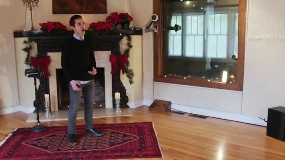 A young adult male in a living room setting with holiday decorations during daytime. He wears a black sweater over a white collared shirt and dark gray jeans.