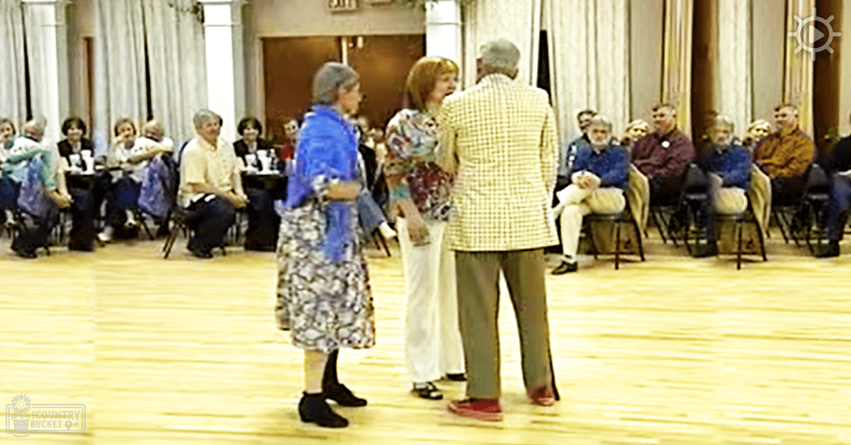 An older adult male singer dancing with a group of older adults in an indoor hall during daytime.
