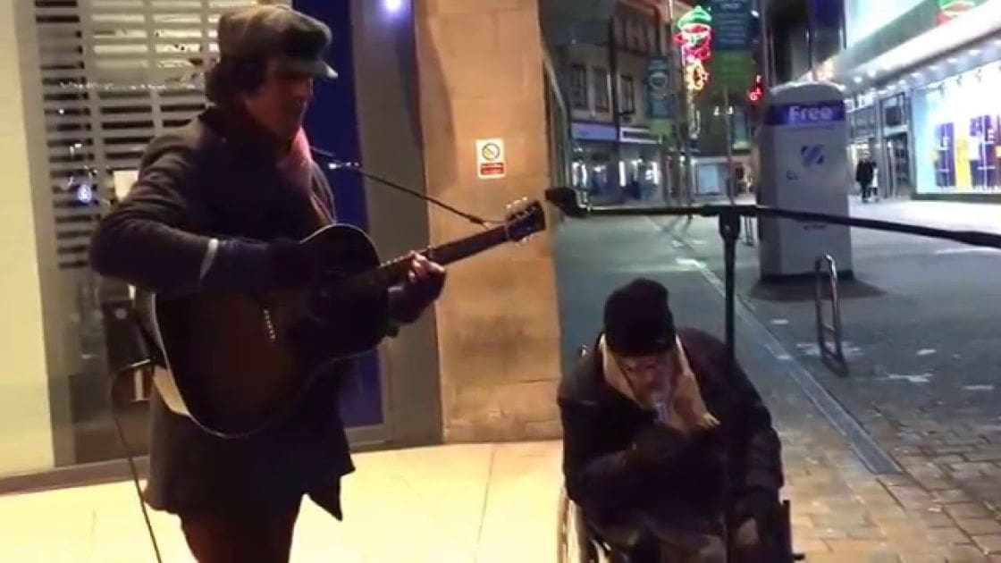 Adult male street performer playing guitar at night in an urban setting with illuminated signs and buildings in the background.