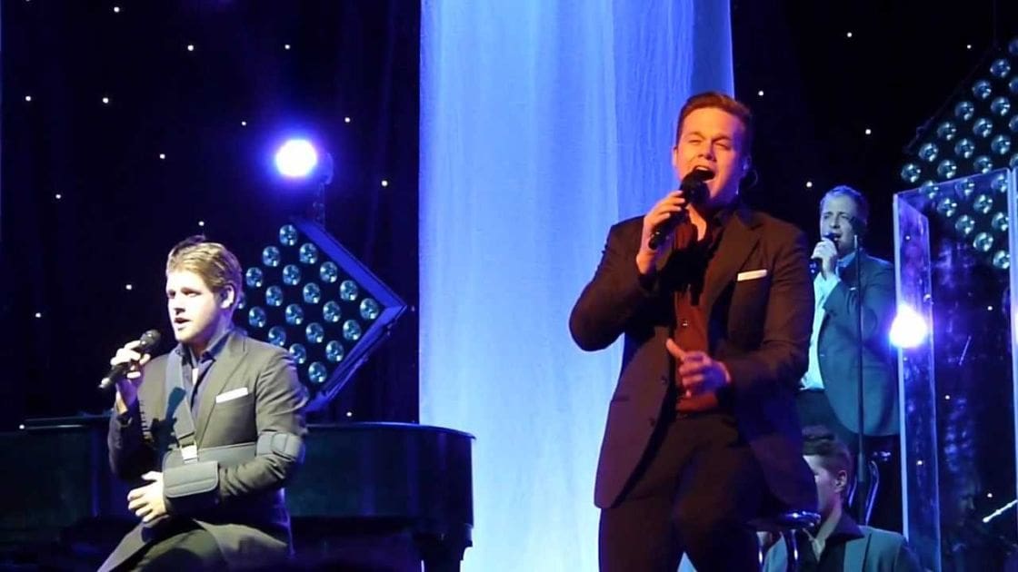 A musical performance on an indoor stage with a black grand piano, decorative lights, and blue tones in the background.