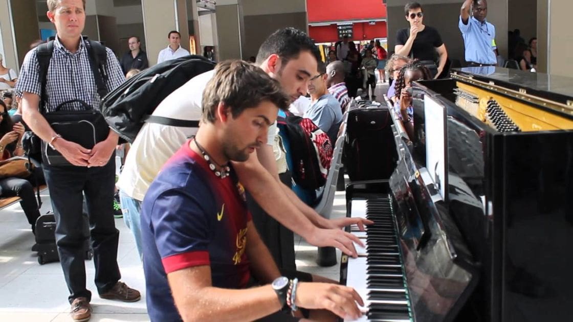 A young adult male playing a piano in an outdoor public space with various adults around him.