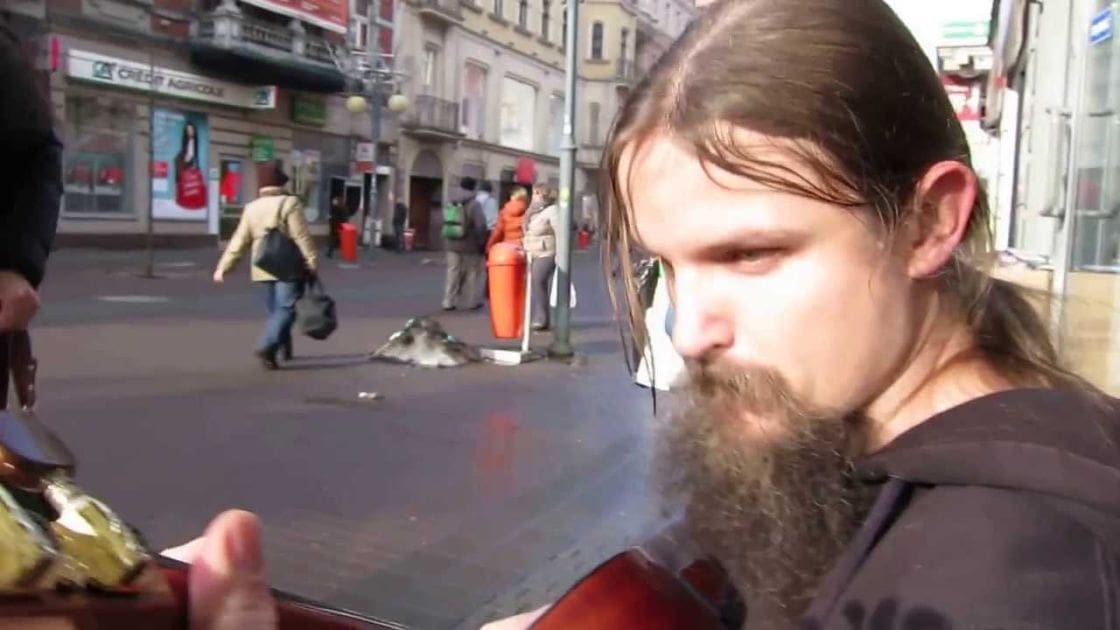 Adult male playing a stringed instrument on an urban street, surrounded by pedestrians and city buildings; daytime scene with various colors and shadows.