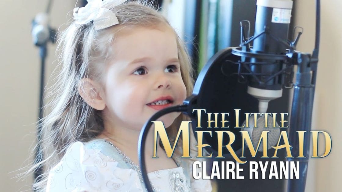 A toddler in a home recording studio, wearing a white garment with swirl patterns and lace details at the hem, performing or recording 'The Little Mermaid' by Claire Ryann.