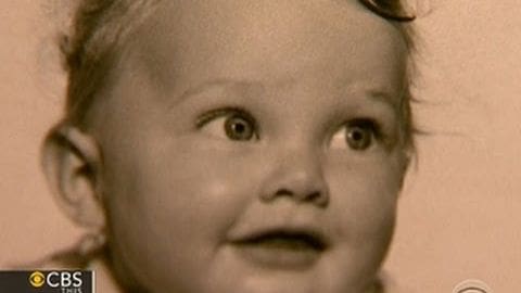 A toddler girl wearing a light-colored garment with a high collar in an indoor setting, evenly lit.