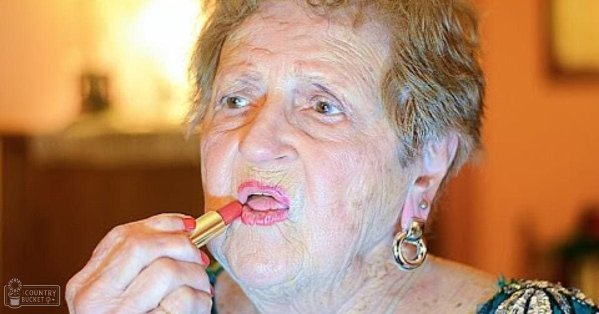 An older adult female wearing a dark green sleeveless garment with embroidered shoulders, applying lipstick indoors near furniture under natural light.