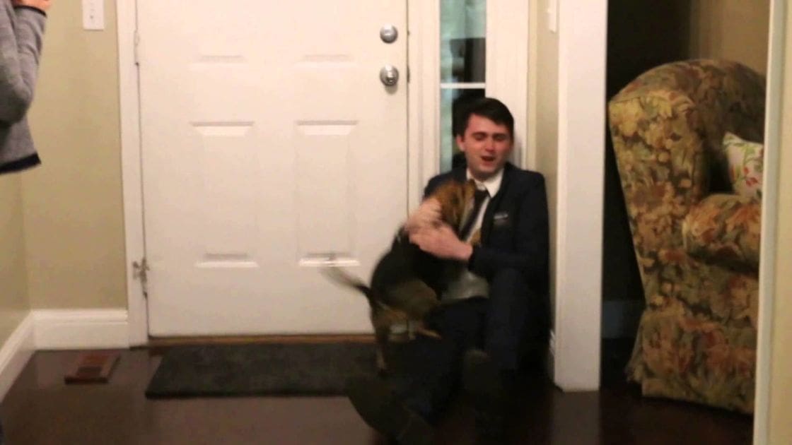 A young adult male holds a tricolor Beagle dog in front of a door inside a home, with floral-patterned furniture and a dark mat on the floor.