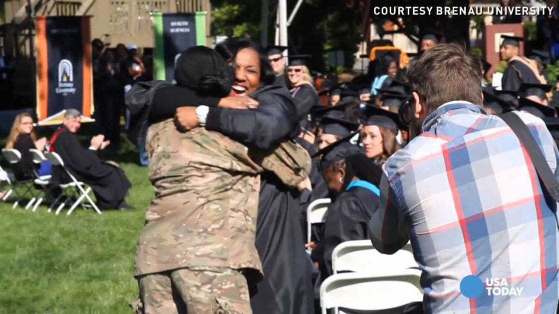 Two graduates, one in camouflage military uniform and another in black graduation gown, embrace at an outdoor Brenau University graduation ceremony under sunny skies with lush green grass.