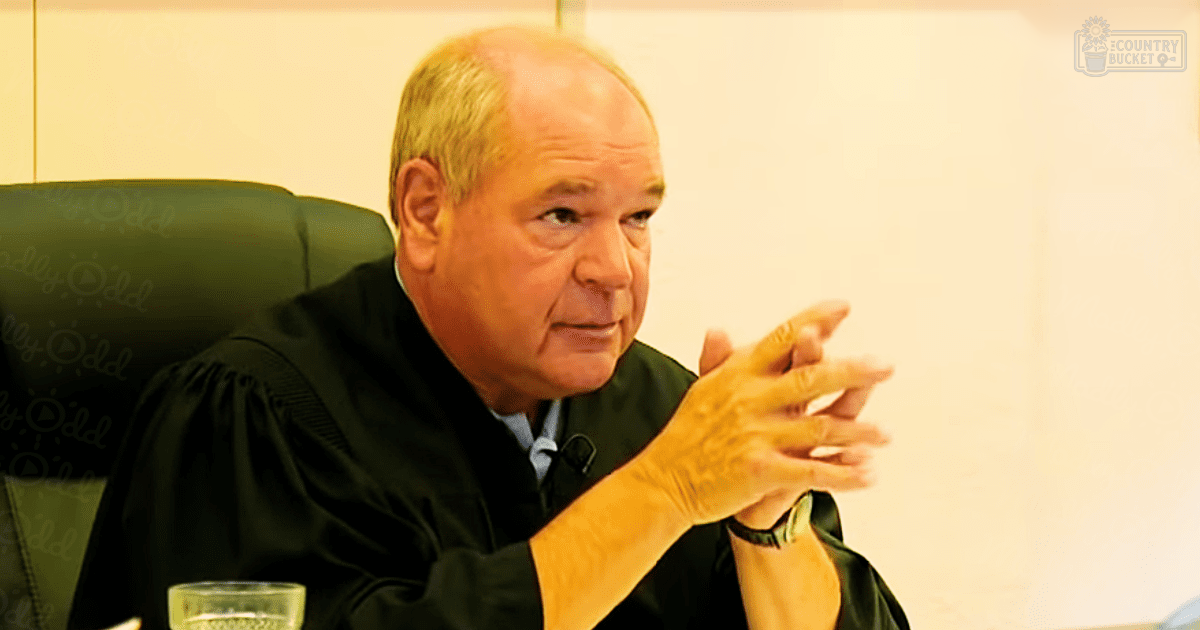 An older adult male judge wearing a black judicial robe and white shirt, seated on a green leather chair with 'Odd' repeated on it in what appears to be a courtroom setting. Natural light suggests daytime.