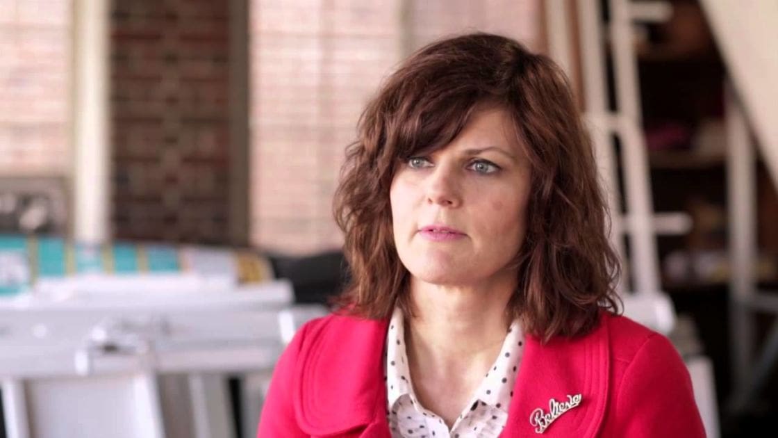 A middle-aged woman in a white polka-dotted blouse with a bright pink cardigan, seated indoors against a brick wall backdrop.
