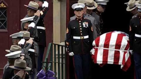 A solemn military funeral ceremony at an American military base with uniformed individuals in dark blue dress uniforms, light brown hats, and a casket draped with an American flag under soft daylight.